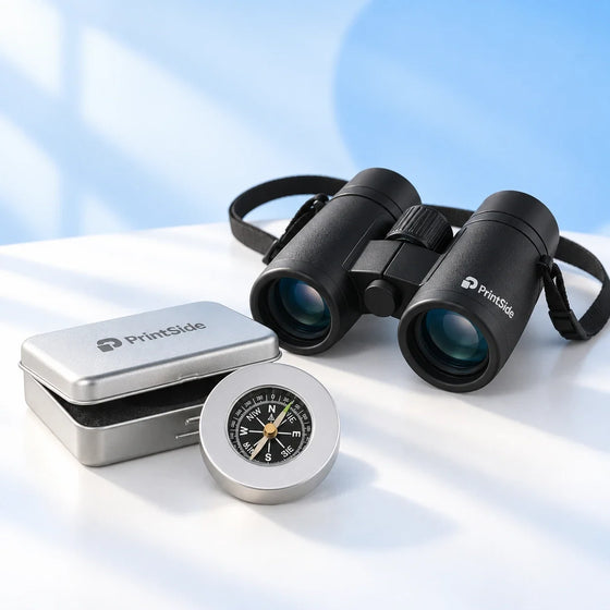Black binoculars beside a silver compass and metal tin with PrintSide logos.
