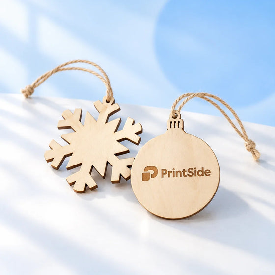 Snöflinga i trä och runt hängande ornament med graverad PrintSide-logotyp.